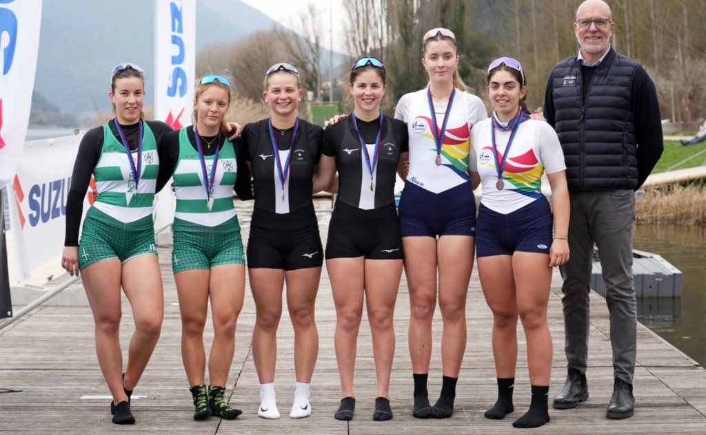 A group of six young female athletes in rowing uniforms standing on a dock, smiling and celebrating with medals around their necks. A man in a vest stands beside them.