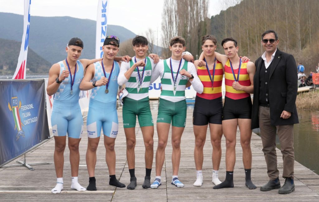 A group of six young male rowers, wearing medals and different athletic uniforms, stand together on a dock by a river, smiling for the camera. A man in a suit stands to the right.