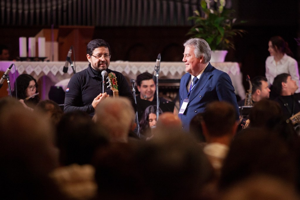 A man speaking into a microphone while holding a guitar, with another man smiling beside him. A musical group is visible in the background, with audience members in front.