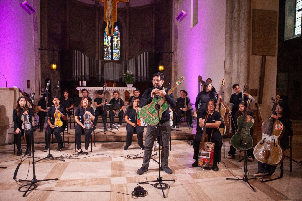 A music ensemble performing in a church, featuring various musicians with string instruments and microphones, under colorful lighting.