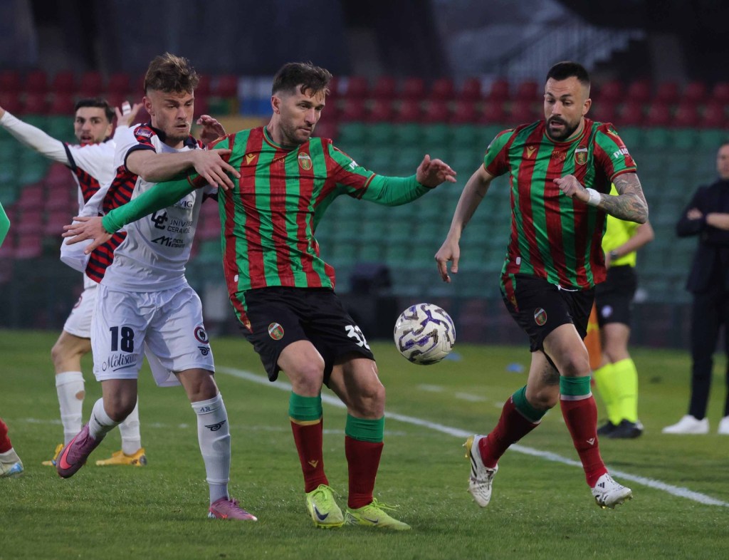 Two soccer players in green and red striped jerseys compete for the ball, while a third player in a white jersey tries to intervene in a dynamic play during a match.