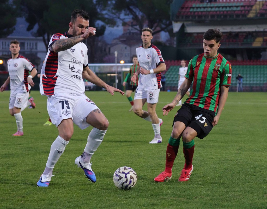 Two soccer players are competing for the ball on a grassy field during a match. One player in a white jersey with red and black stripes is about to take a shot, while the other in a green and red striped jersey is positioned to intercept.