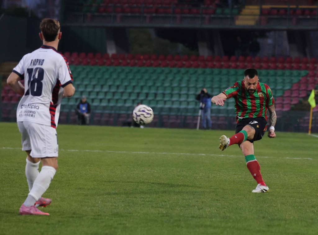 A soccer player in a red and green striped jersey strikes a ball on the field, while a player in a white jersey with the number 18 watches from a distance.