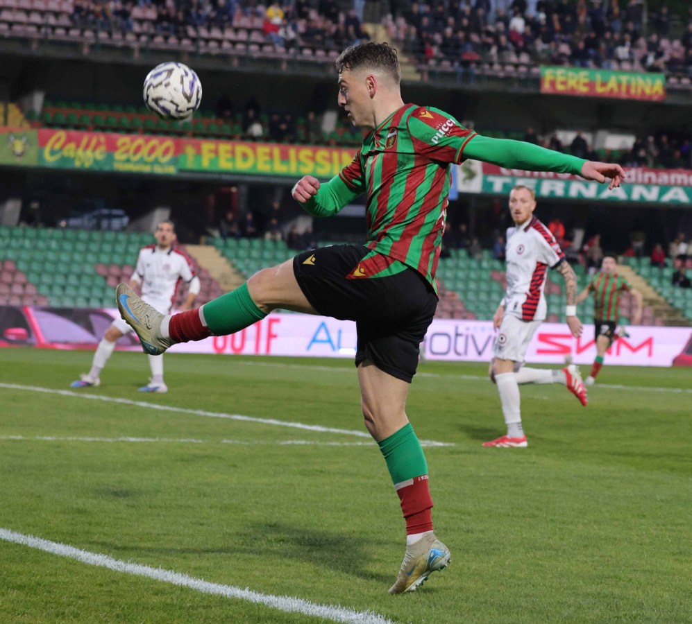 A soccer player in a striped green and red jersey kicks the ball while another player in a white jersey watches. The scene takes place on a grassy soccer field with spectators in the background.