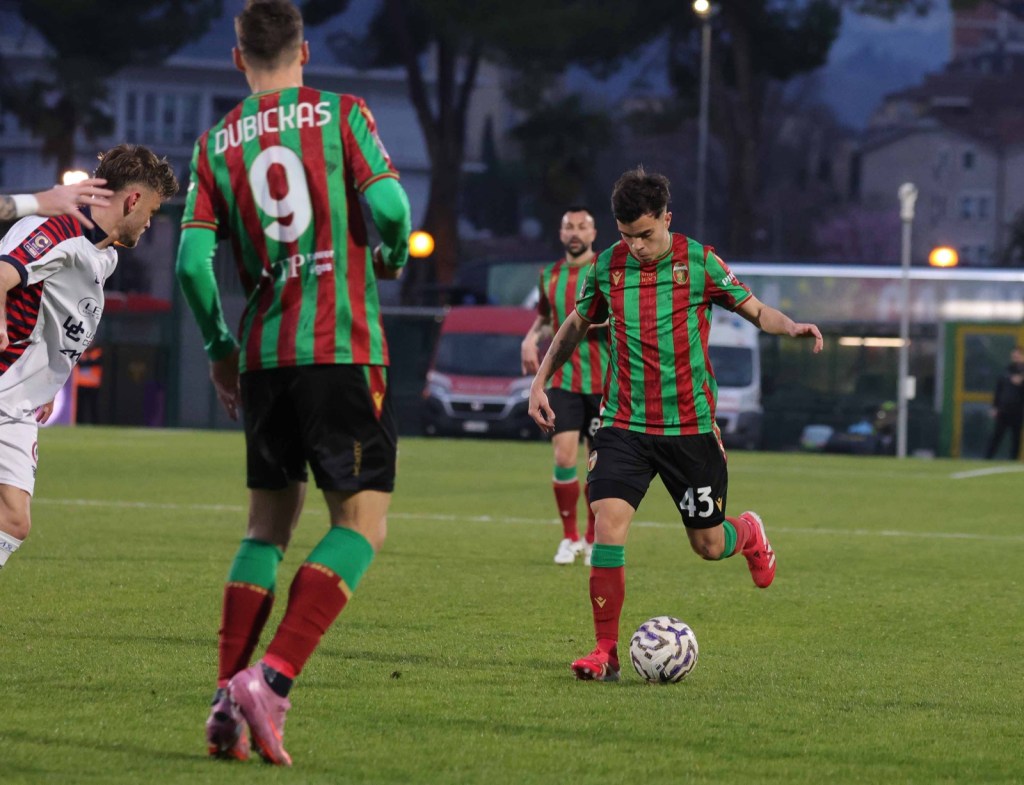 A soccer player in a red and green striped jersey dribbles the ball on the field while two opposing players watch.
