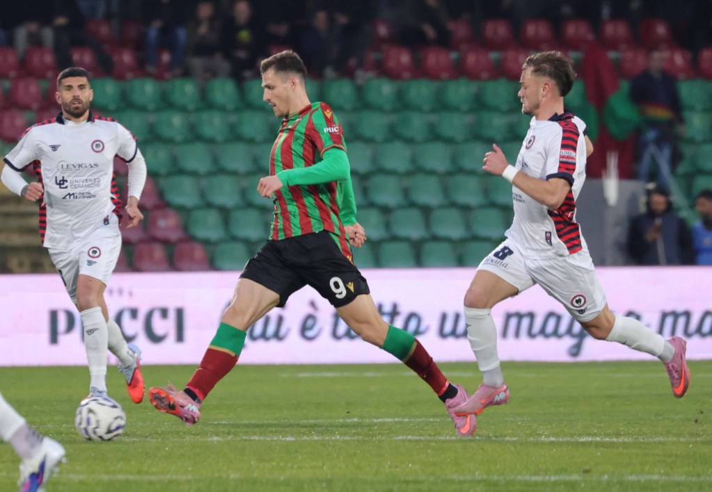A football player in a green and red striped jersey attempts to kick the ball while two opposing players in white jerseys try to defend.