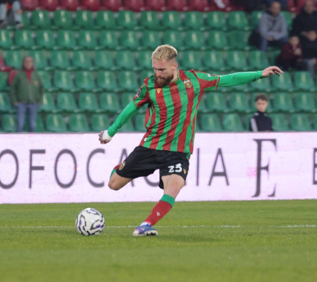 A soccer player in a red and green striped jersey is kicking a soccer ball on the field during a match.