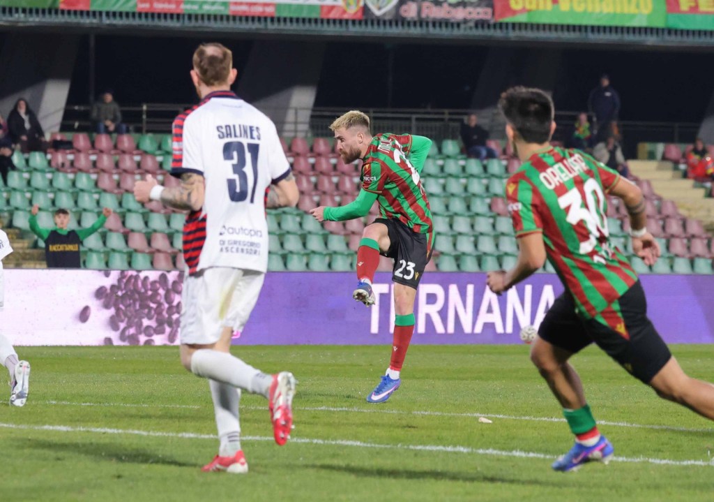 A soccer player in a green and red kit jumps in celebration after scoring a goal, while two teammates and an opposing player look on. The stadium has a colorful backdrop with cheering fans.