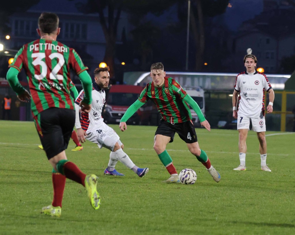 A soccer match in progress with players in colorful uniforms. One player in a red and green jersey is dribbling the ball, while another in a white jersey is positioned nearby. The scene captures the action on a grassy field during an evening game.