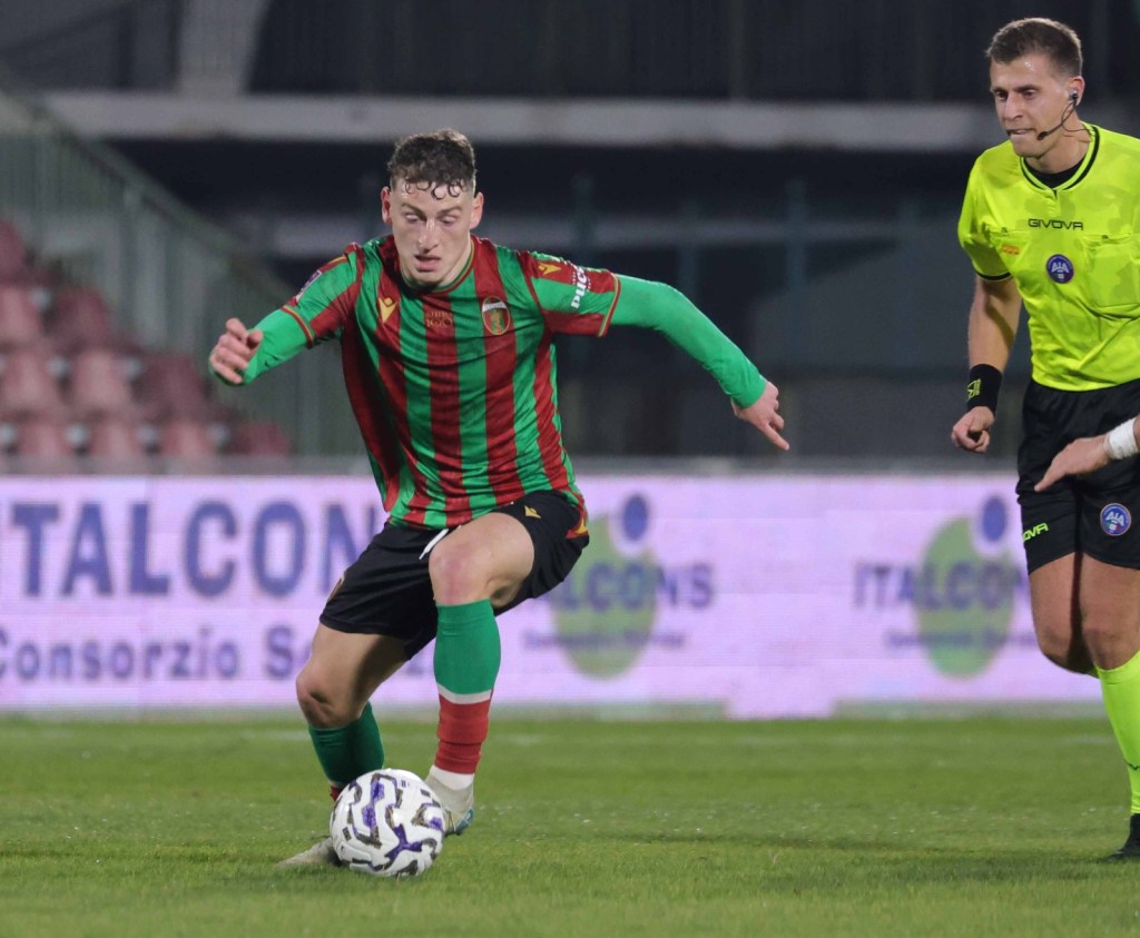 A soccer player in a red and green striped jersey is dribbling the ball on the field, while a referee in a yellow shirt observes the play.