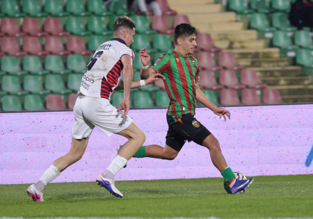 A football match scene showing a player in a green and red striped jersey sprinting with the ball, while an opposing player in a white and red jersey is closely following.