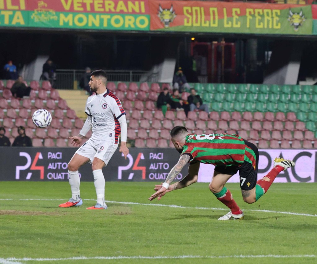 A soccer player in a green and red striped jersey dives to save a ball, while another player in a white kit watches. The scene takes place on a football field with empty stands in the background.