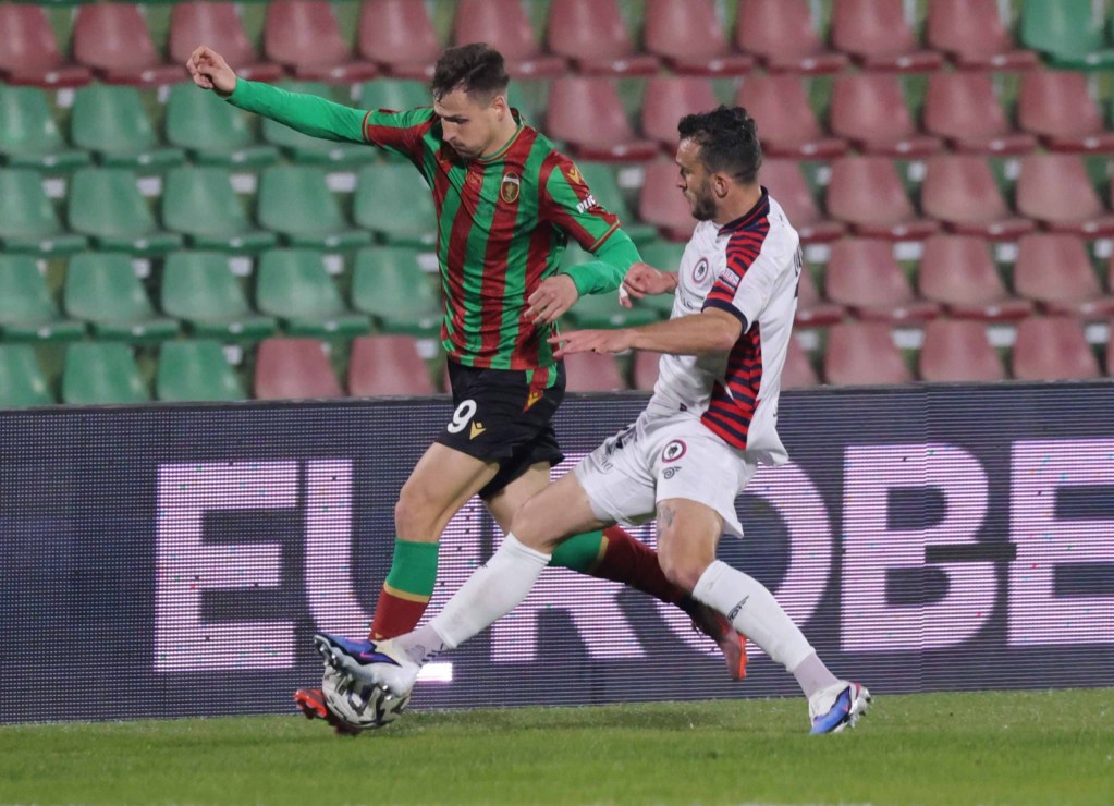 A football match action shot showing a player in a green and red striped uniform being challenged by an opponent in a white and red uniform, with the background featuring empty stadium seats.