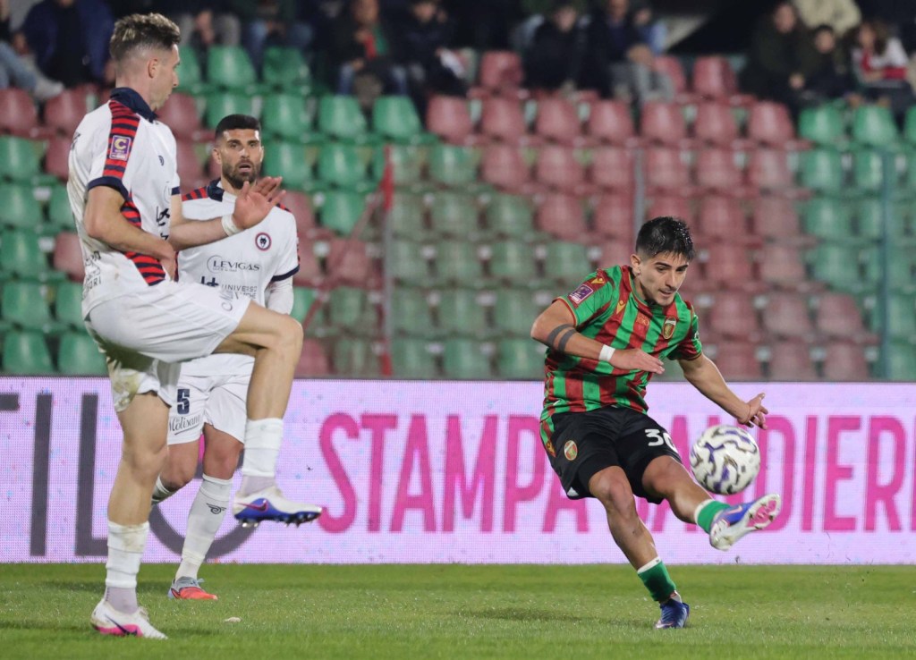 A soccer player in a striped green and red jersey prepares to kick the ball while two opponents in white uniforms look on during a match.