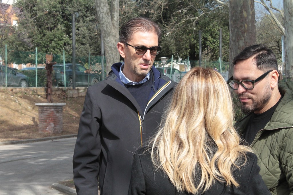 Three people engaged in conversation outdoors, with green trees and parked cars in the background. One person is wearing sunglasses and a dark jacket, another has long blonde hair, and the third is wearing glasses and a green jacket.