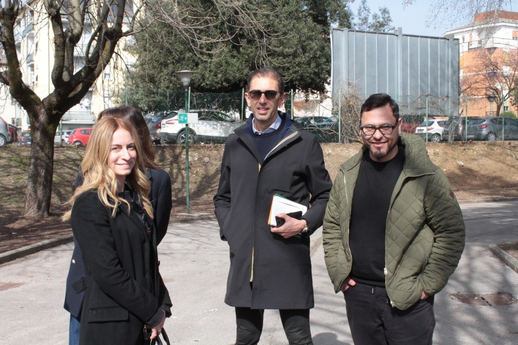 A group of four people standing outdoors in a park. Two women and two men are smiling and engaging with each other. The background features trees and parked cars.