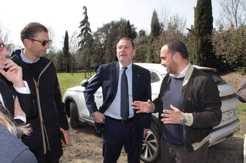 Group of four men engaged in conversation outdoors, with a white vehicle parked in the background and trees surrounding the area.