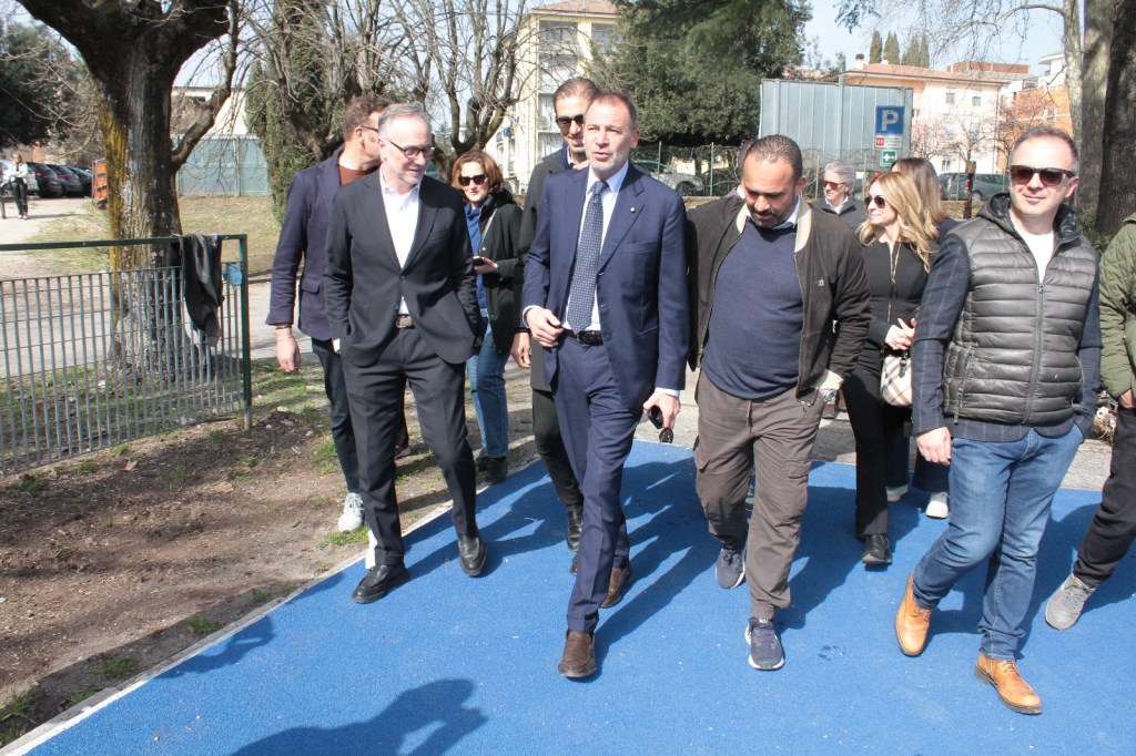 A group of people walking together on a blue pathway in a park, with trees in the background and a parking area visible.