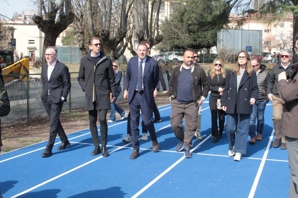 A group of individuals walking on a blue track in a park, with trees and a construction vehicle in the background.