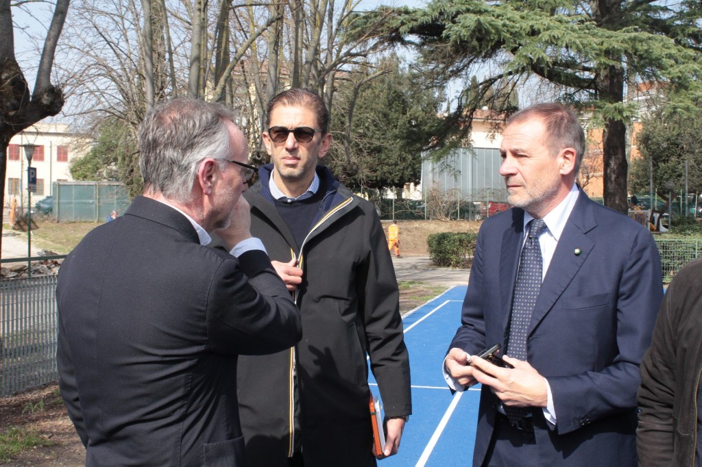 Three men are engaged in conversation outdoors near a blue running track, surrounded by trees and greenery.