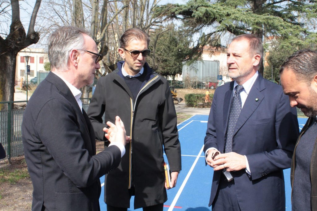 Four men engaged in conversation outdoors near a blue sports court, surrounded by trees.