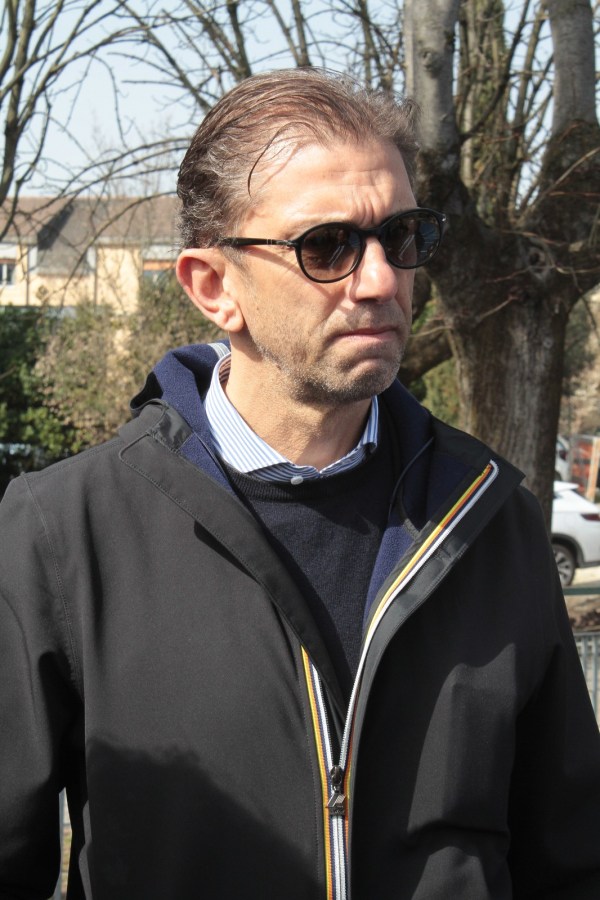 A man wearing sunglasses and a black jacket, looking serious outdoors with trees in the background.