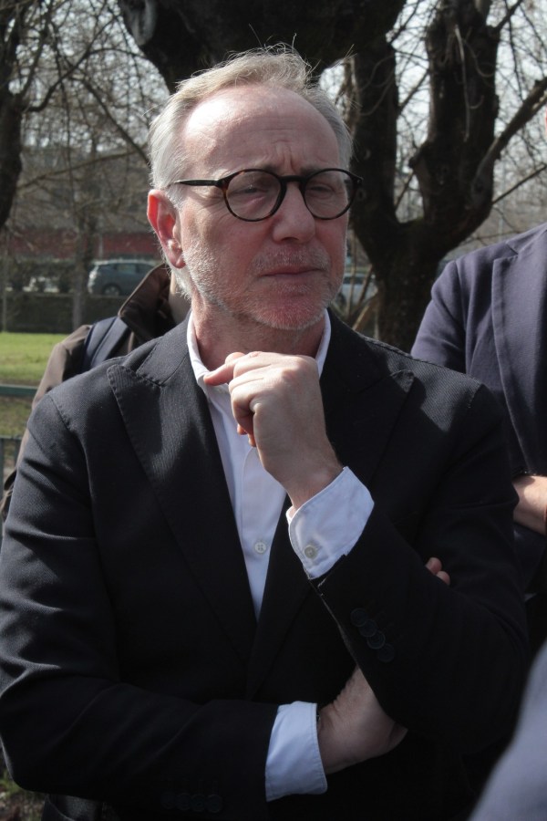A thoughtful man in a black suit and glasses, standing outdoors with his hand on his chin, surrounded by trees.