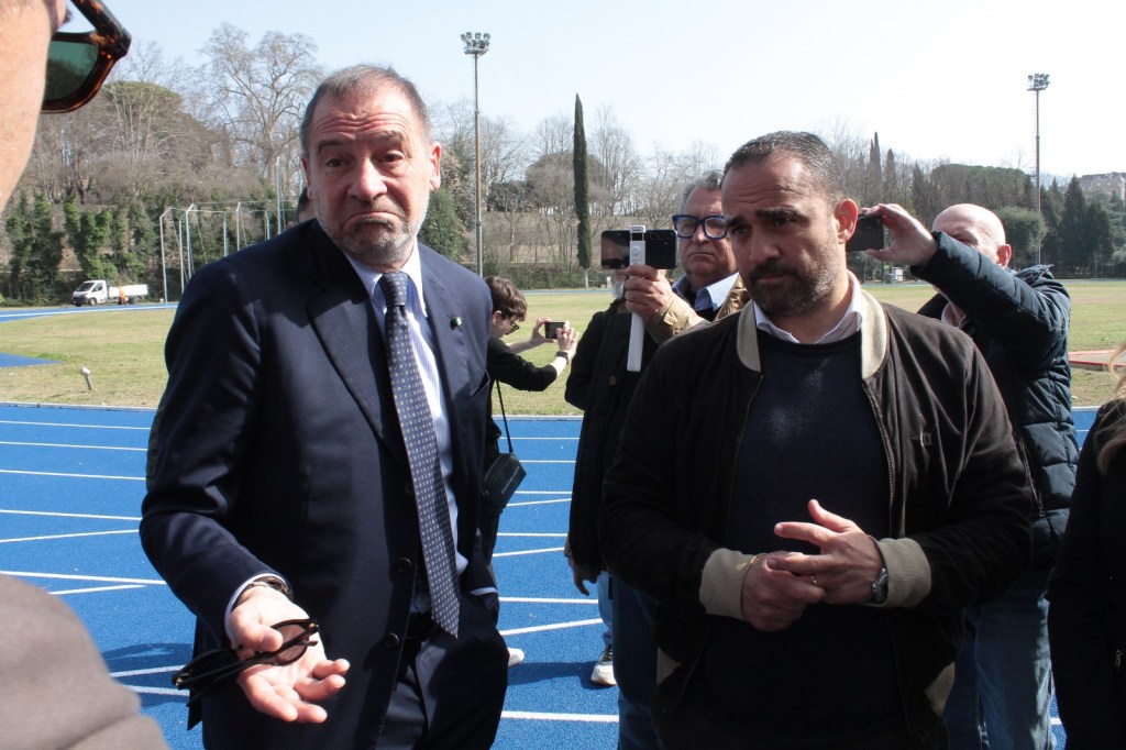 Two men speaking in a sports field, surrounded by a crowd with visible smartphones. One man looks concerned while the other appears serious.