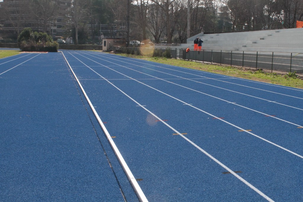 A view of a blue running track with white lane markings, surrounded by greenery and a seating area in the background.