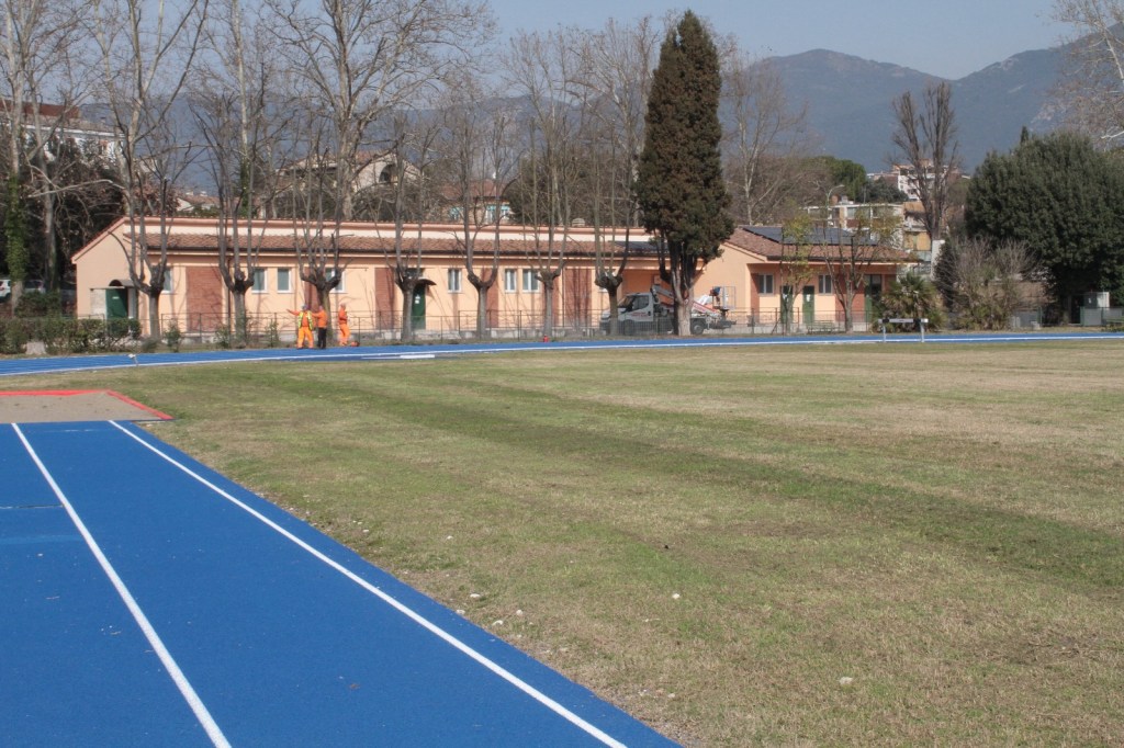 A blue athletic track leading to a sports facility surrounded by trees and grass, with a few workers dressed in orange visible near the building.