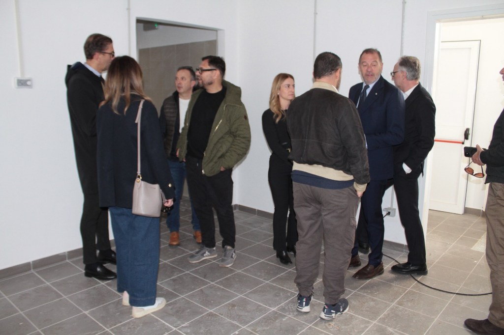A group of people engaged in conversation inside a newly constructed room with gray tiled flooring and white walls.