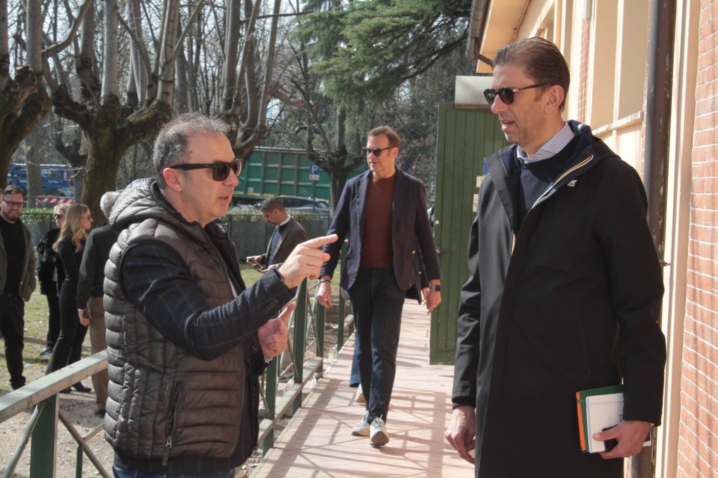 Two men in outerwear converse outdoors while a third man walks past them in a park setting. The trees are bare, indicating early spring.