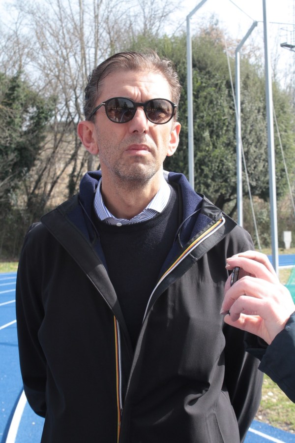 A man wearing sunglasses and a black jacket stands outdoors with a serious expression, while another person's hand is visible holding a device near him, against a backdrop of trees and a sports track.