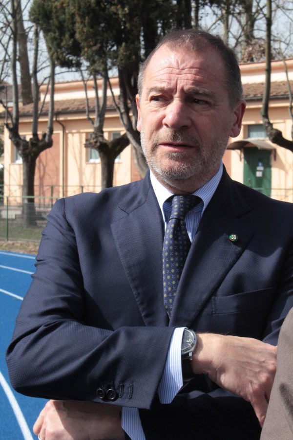 A man in a suit with crossed arms, standing outdoors near a sports field, with trees and a building in the background.