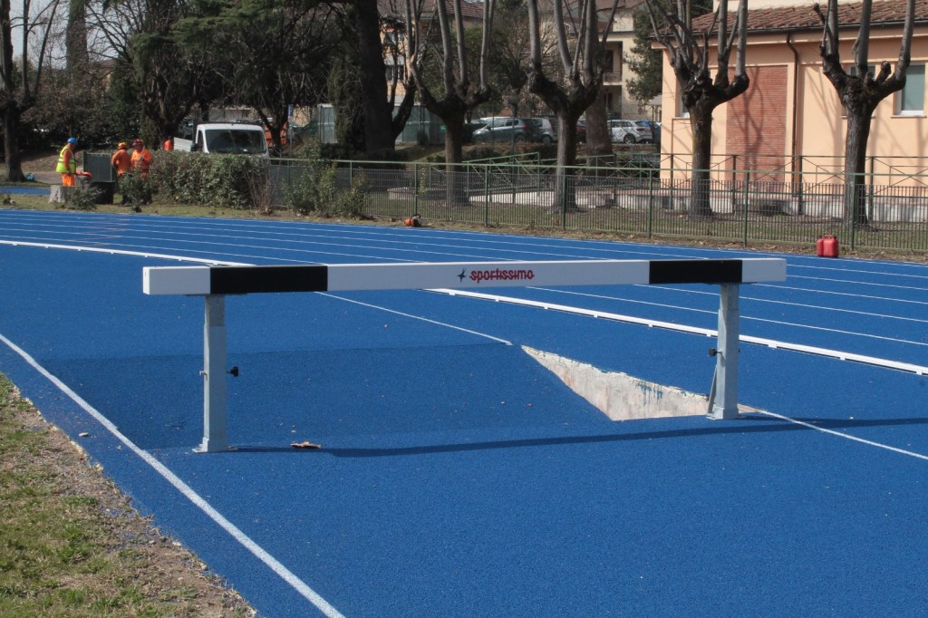 A hurdle positioned on a blue athletic track, with a water jump pit visible in the background. Workers in bright vests can be seen in the distance, attending to the surrounding area.