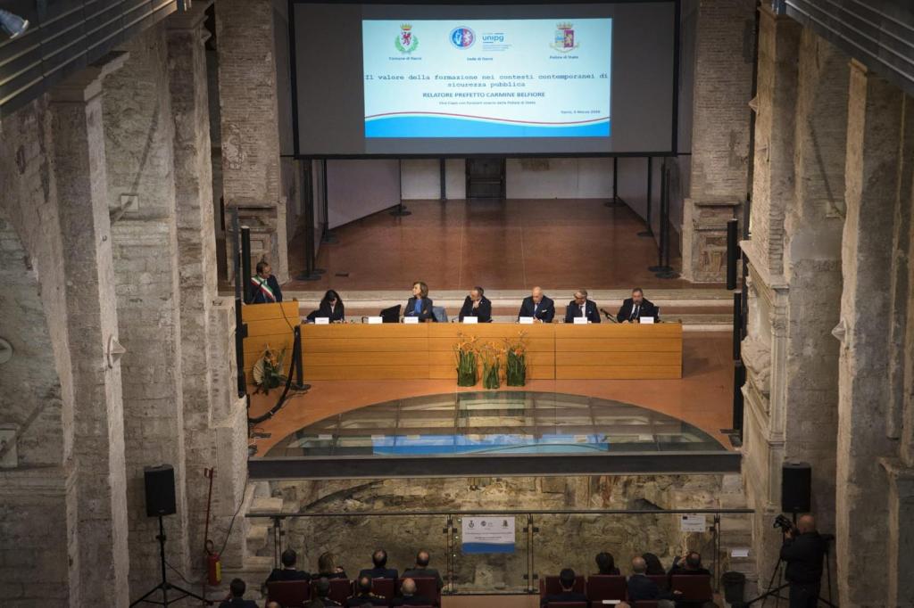 A panel discussion taking place in a historic venue, featuring several speakers seated at a long table, with a large screen displaying information in the background.