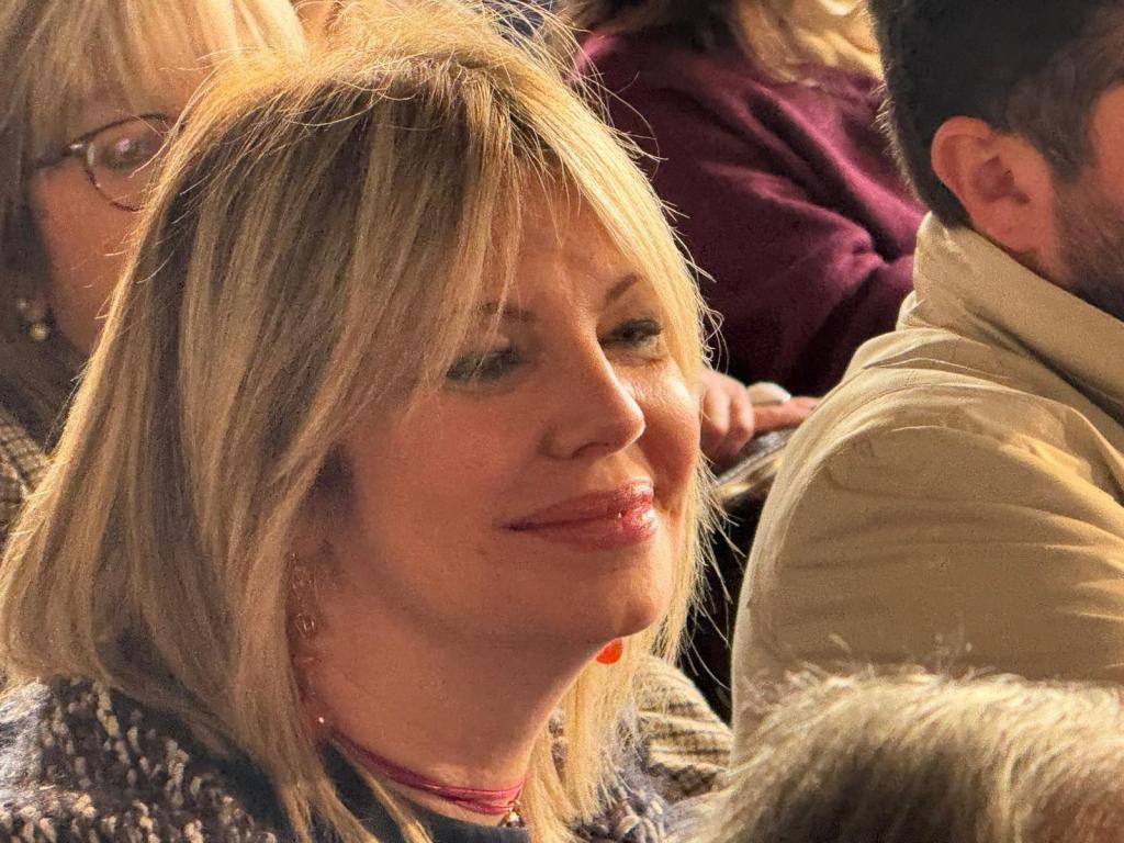 A woman with blonde hair and a joyful expression, sitting in a crowd of people, wearing a cozy outfit.