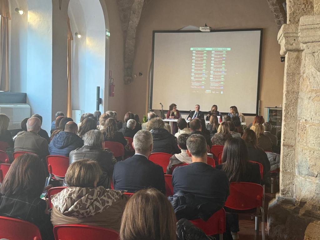 A conference or panel discussion taking place in a spacious venue, with a seated audience facing a panel of speakers at a table. A projector screen displaying text is visible behind the speakers, with soft lighting and a historic architectural backdrop.