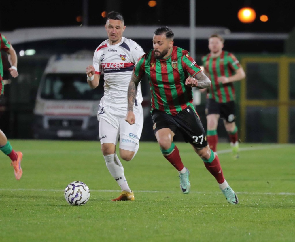 Two soccer players are competing for the ball on a grass field, with one player in a green and red striped jersey chasing while the other is in a white jersey.