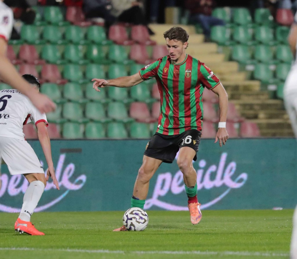 A soccer player in a red and green striped jersey dribbles a ball on the field during a match, with another player in a white jersey nearby.