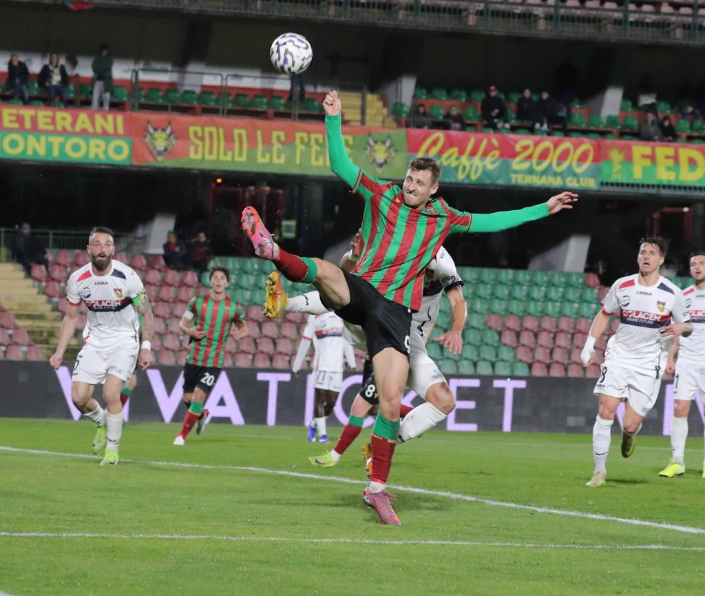 A soccer player wearing a red and green striped jersey performs a kick in mid-air, while other players in white jerseys are positioned in the background.