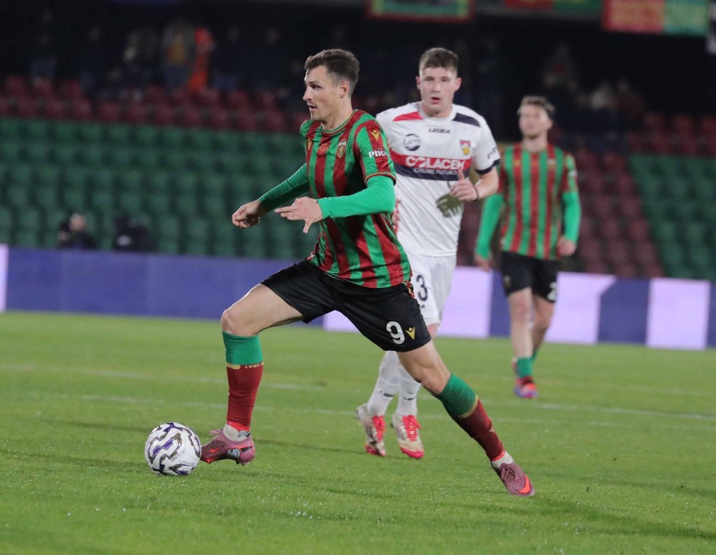 A football player in a red and green striped jersey dribbles the ball on the field during a match, with teammates and opponents in the background.