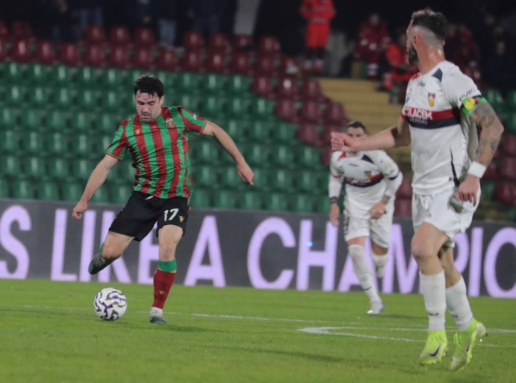 A football player wearing a red and green striped jersey with the number 17 is in motion, preparing to kick the ball on a green pitch during a match. An opposing player in a white uniform is seen nearby, along with other players in the background.