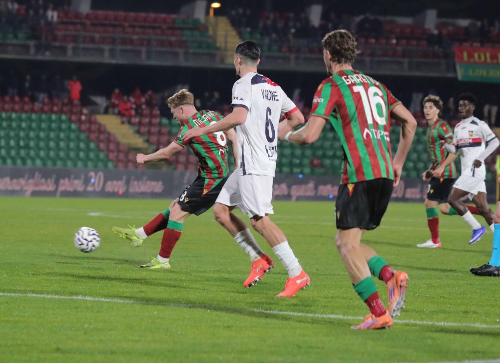 A football match scene showing players in action on the field. One player in green and red is about to kick the ball, while two other players in white and black are positioned nearby. The stadium is filled with spectators in the background.