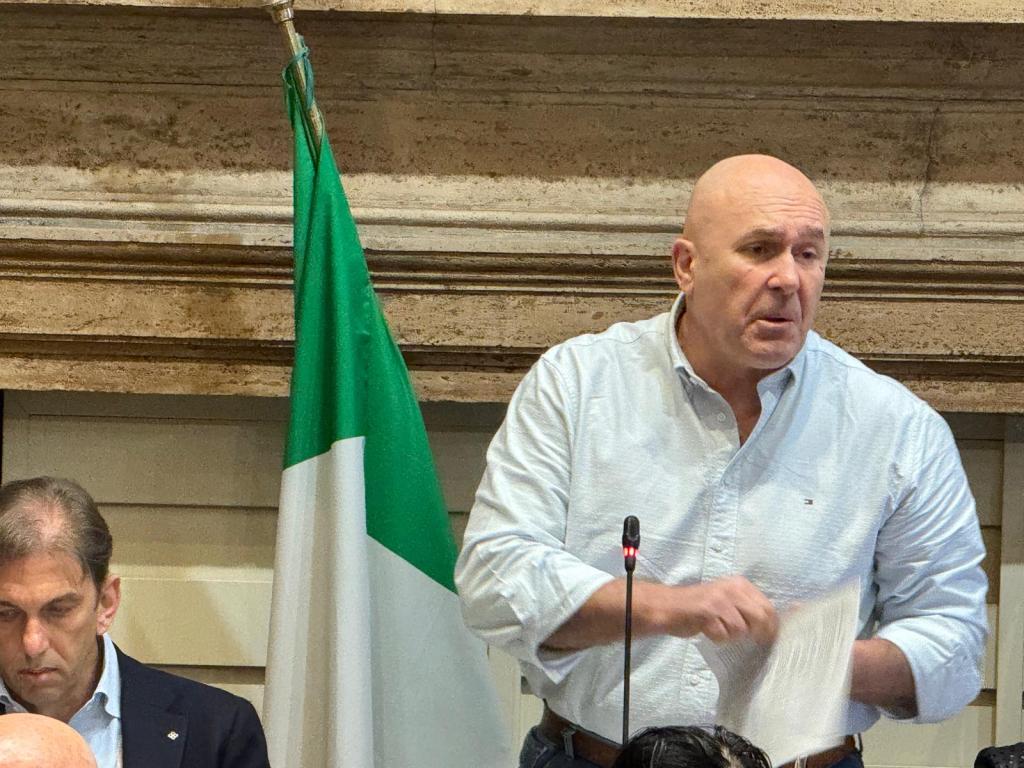 A man speaking at a podium with a microphone, gesturing while holding a piece of paper, with a green and white flag in the background.