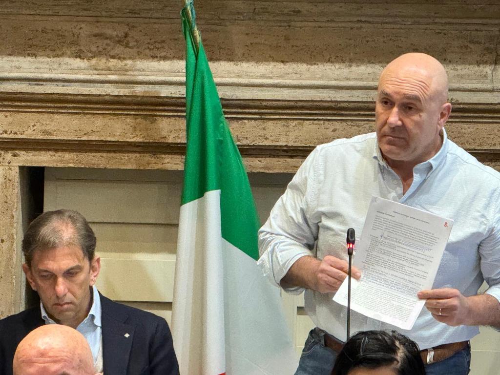 A man holding a document stands while speaking at a podium, with an Italian flag visible in the background. Another man seated below appears to be listening attentively.