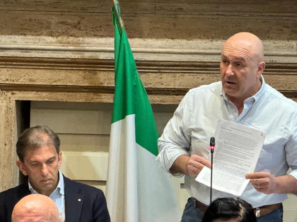 A man holding a paper speaks actively at a podium while another man sits thoughtfully beside him, with an Italian flag in the background.