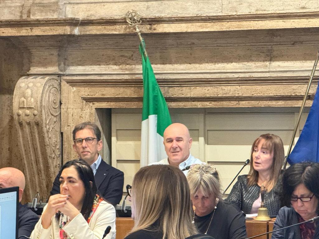 A group of people seated at a conference table, with an Italian flag and a blue flag visible in the background. Several individuals are engaged in discussion, while one person appears to be speaking.
