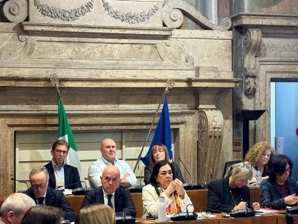 A group of individuals seated at a meeting table, with Italian and European flags in the background, inside a historic building.