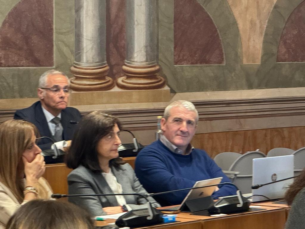 A group of people sitting at a conference table, with a decorative background. One man with glasses is in the back, while two women and another man are in the foreground, engaged in discussion.